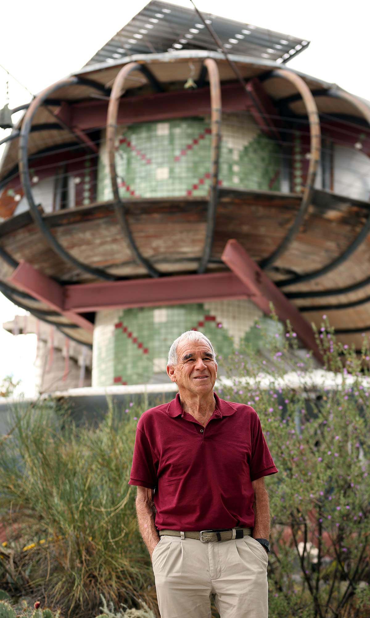 Bart Prince wearing a red shirt and khaki pants, standing in front of his home which looks like a spaceship and smiling.