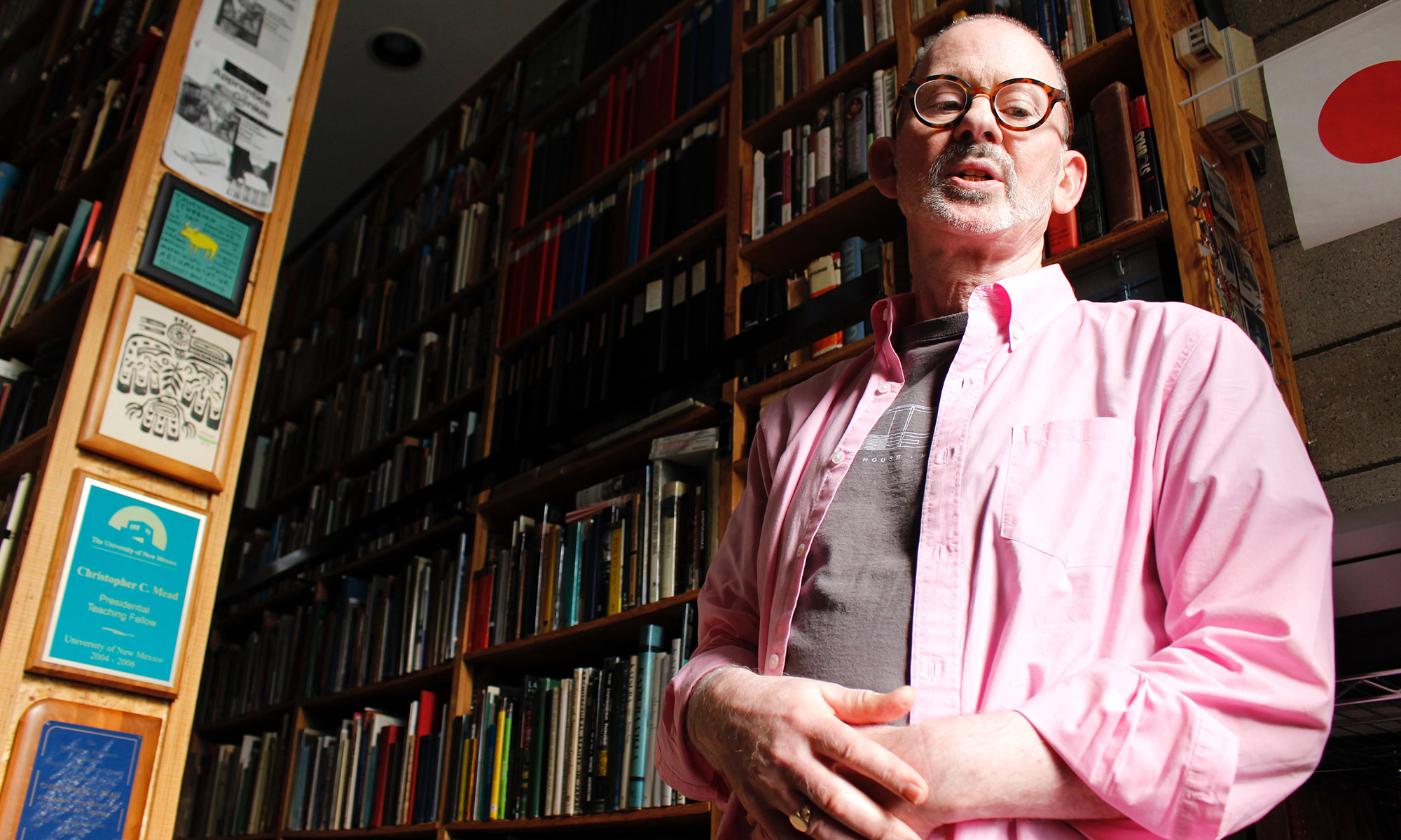Portrait of Christopher Mead, wearing a pink shirt and standing in front of a huge bookshelf completely full of books