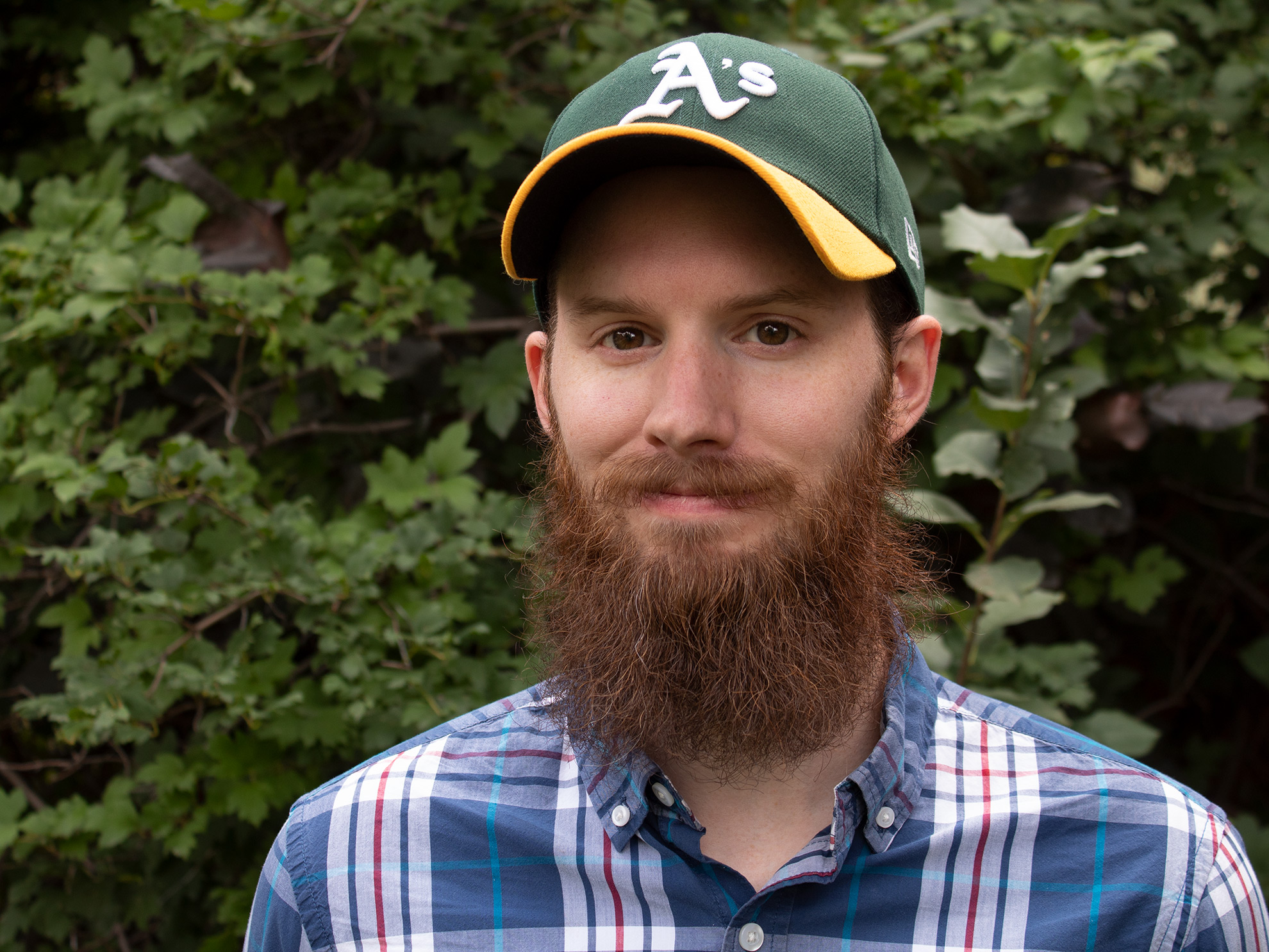 Ron Stauffer with a large beard, smiling at the camera and wearing an Oakland Athletic's ballcap standing in front of a bush.